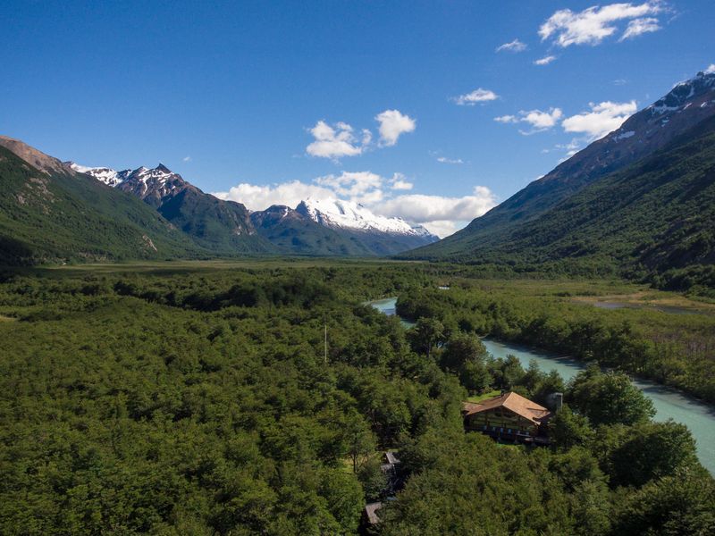 Snow-capped mountain valley with turquoise river, dense forest, and rustic lodge near Laguna Cóndor in Patagonia.
