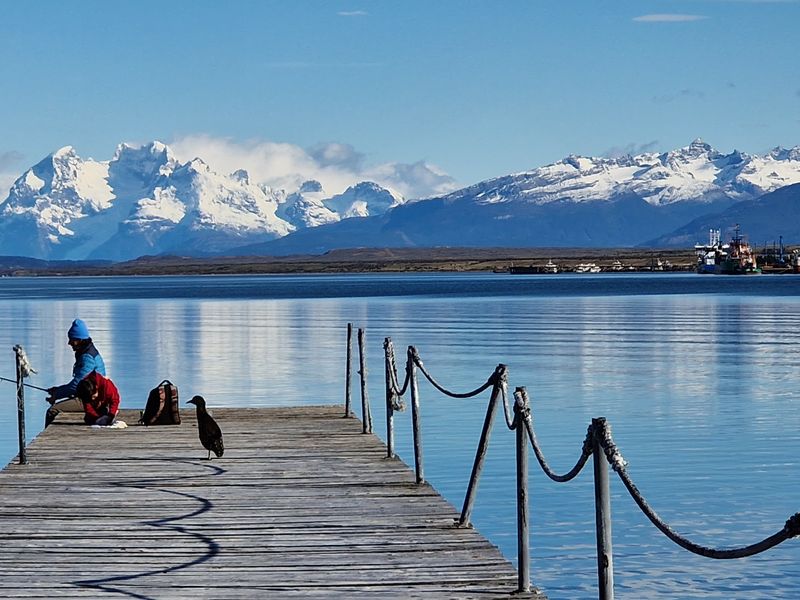 Wooden dock at Puerto Natales with snow-capped Patagonian mountains reflected in pristine blue waters.