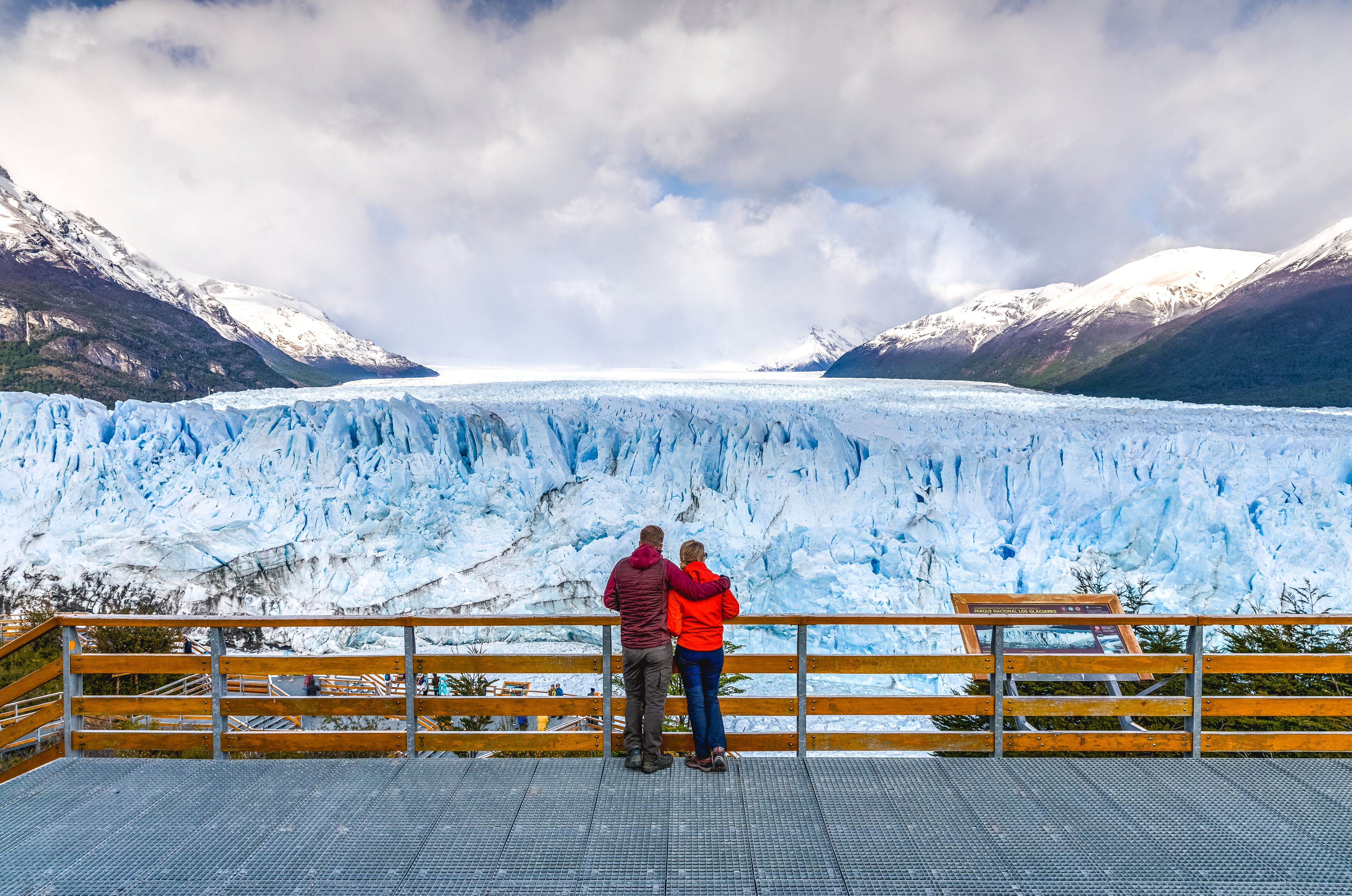 Two people on a viewing platform look at a vast blue glacier, framed by snowy mountains under a cloudy sky.