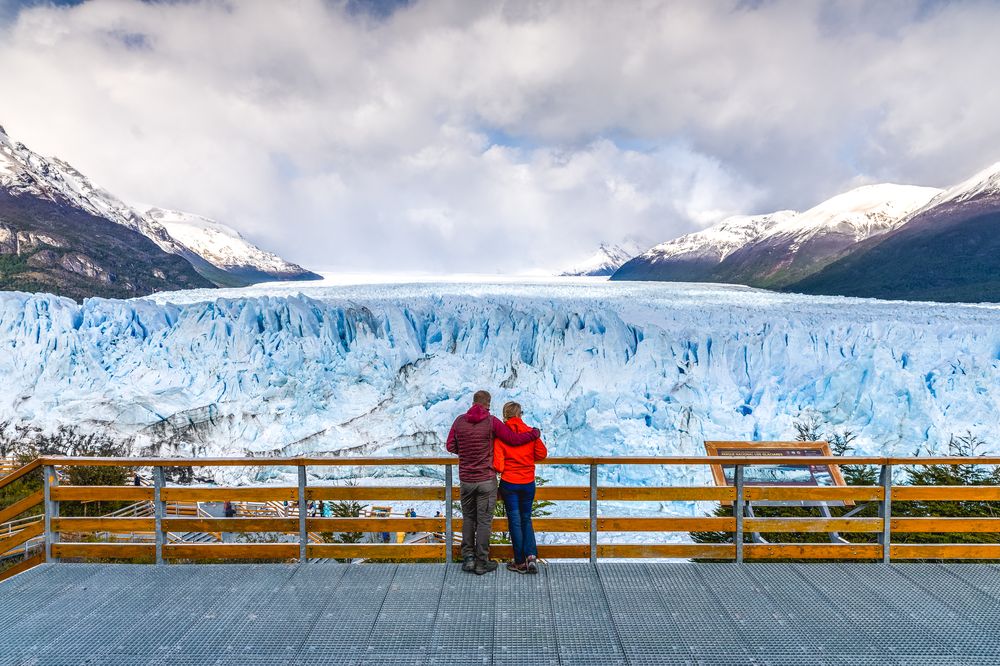 Two people on a viewing platform look at a vast blue glacier, framed by snowy mountains under a cloudy sky.