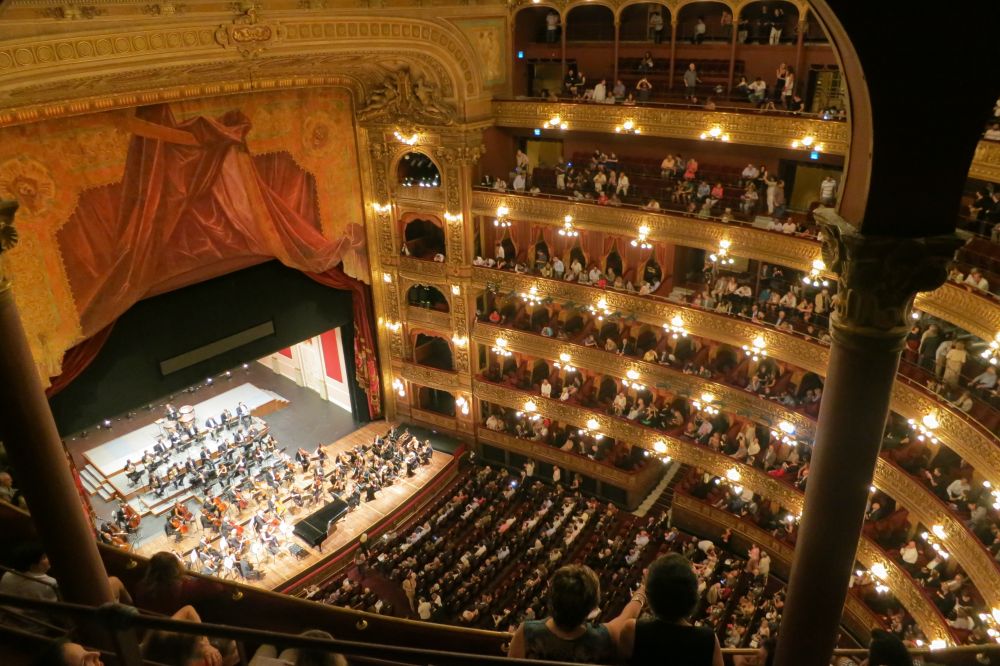 Grand ornate interior of Teatro Colón in Buenos Aires with orchestra performing on illuminated stage below multiple tiers of balconies.