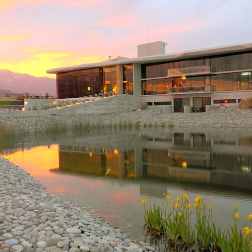 Modern architecture at Casa de Uco reflects in still waters at sunset, with snow-capped Andes mountains in the distance.