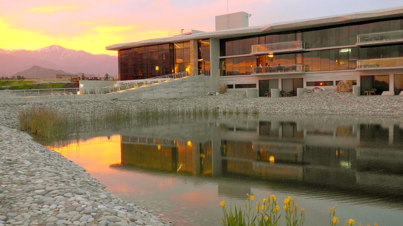 Modern architecture at Casa de Uco reflects in still waters at sunset, with snow-capped Andes mountains in the distance.