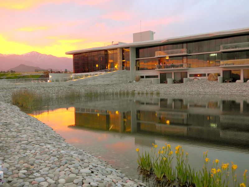 Modern architecture at Casa de Uco reflects in still waters at sunset, with snow-capped Andes mountains in the distance.