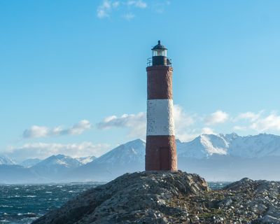Red and white striped lighthouse on rocky outcrop with snow-capped mountains and turbulent waters near Ushuaia, Patagonia.