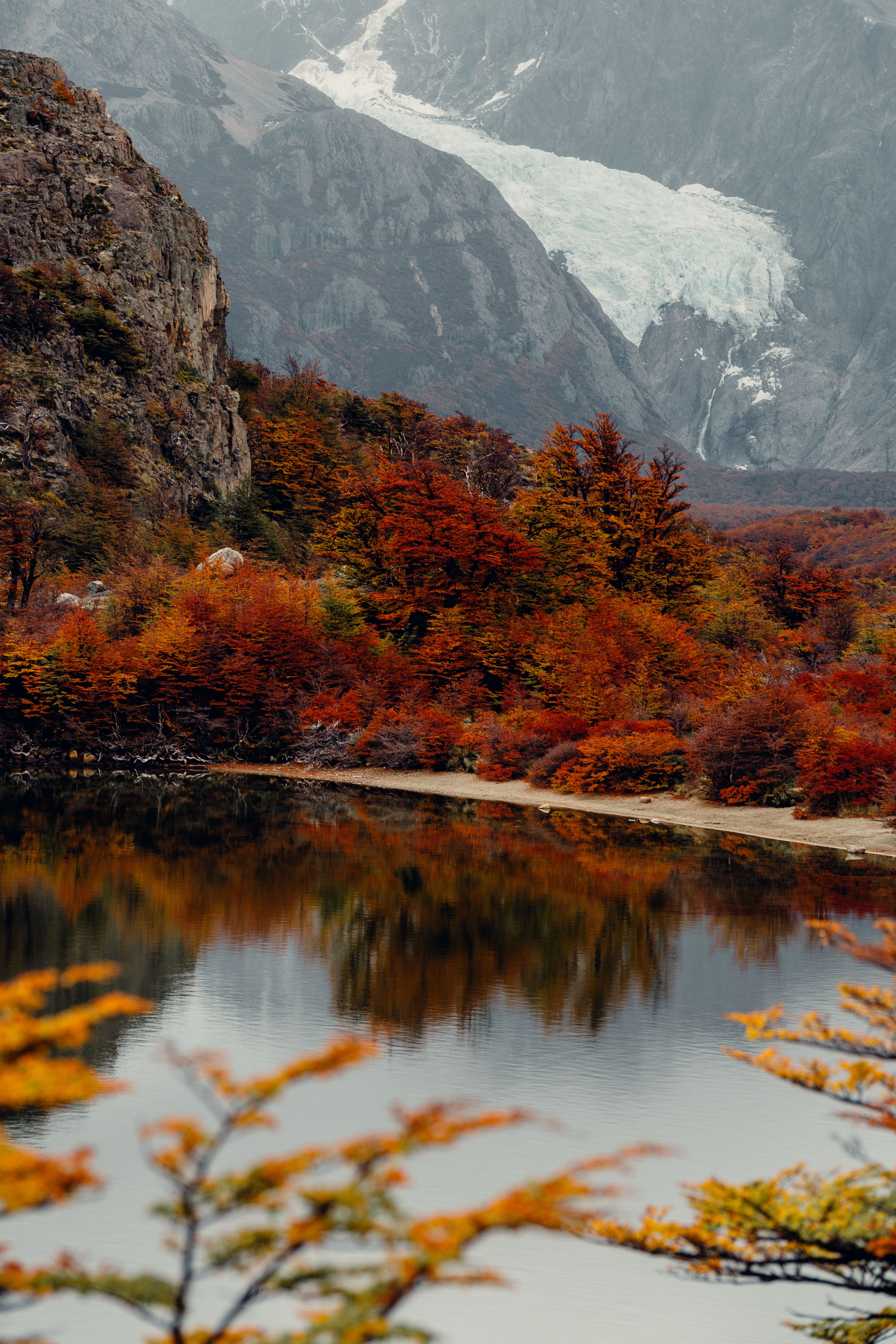 Autumn foliage reflects in a pristine lake near El Chaltén, with a glacier-fed river and snow-capped mountains in the distance