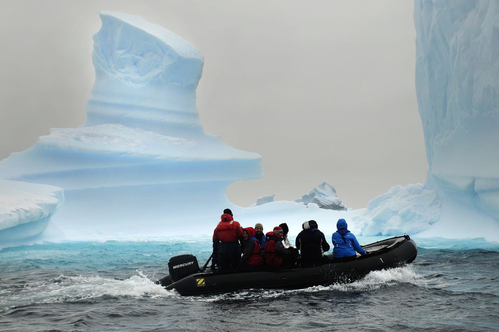 Zodiac boat expedition near massive Antarctic icebergs with tourists in cold-weather gear exploring pristine polar waters.