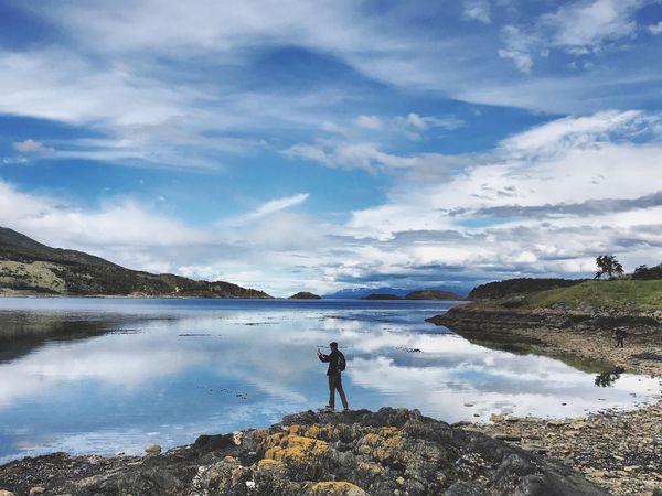 Hiker standing on rocky outcrop overlooking pristine Patagonian lake with mirror-like reflections and mountain peaks under dramatic blue sky.
