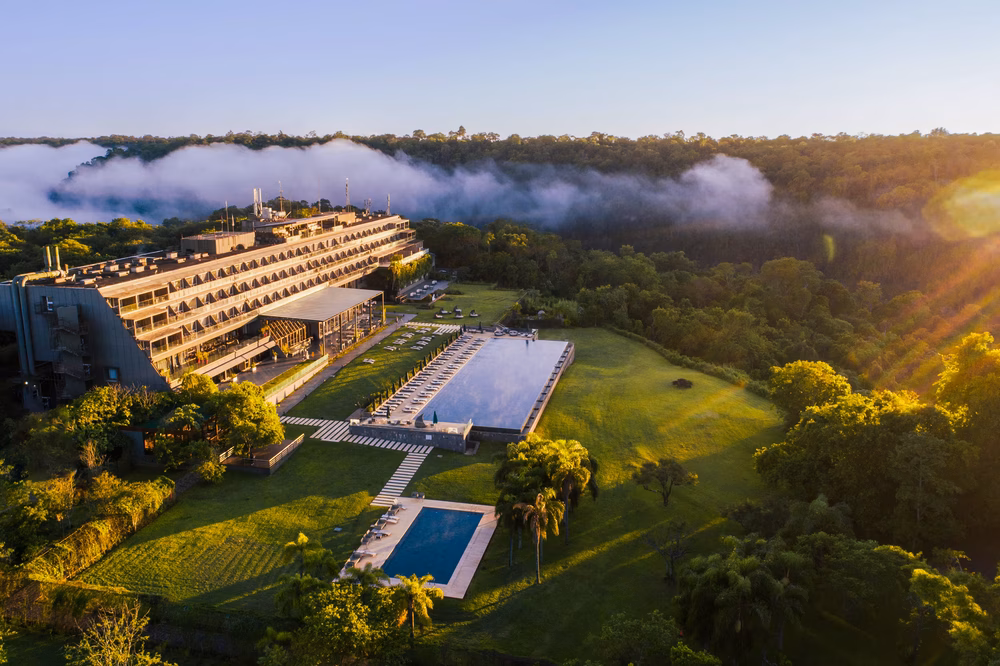 Luxury hotel with modern architecture overlooks lush grounds, swimming pools, and misty Iguazú Falls landscape at sunrise in Argentina.