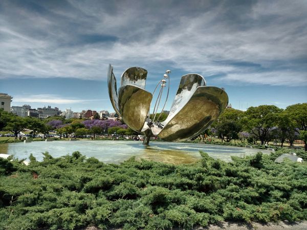 Large metallic flower sculpture beside a lagoon in Buenos Aires with jacaranda trees and urban skyline.