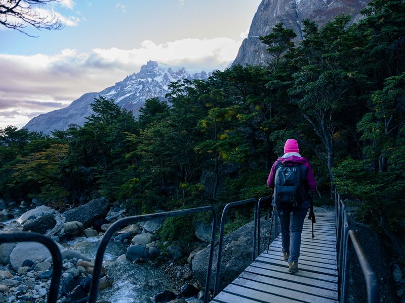 Hiker in pink jacket crossing wooden bridge through lush forest with snow-capped peaks in Valle del Francés, Torres del Paine, Chile.