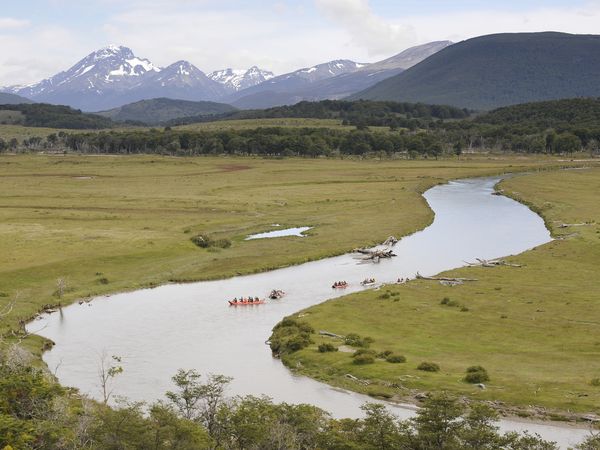 People in red rafts paddle a winding river through a wide green valley, flanked by forests and distant snow-capped peaks in Ushuaia.