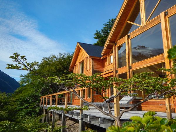 Modern wooden lodge nestled in Patagonian forest with mountain views and expansive glass windows.