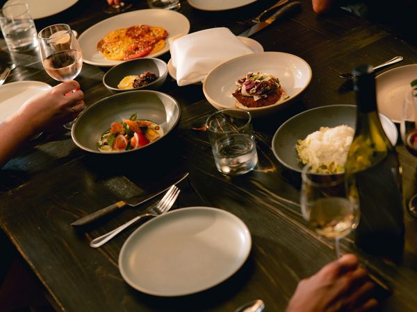 Overhead view of a dinner party: hands, wine glasses, various artfully plated small dishes on a dark wooden table.