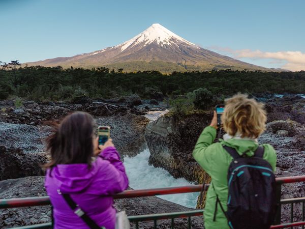 Hikers photograph Villarrica volcano from a turquoise river viewpoint in Chile's Lake District during Cruce Andino trek.