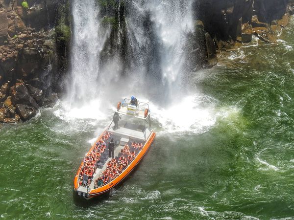 Adventure boat packed with tourists navigates turbulent waters beneath a powerful waterfall in Brazil.