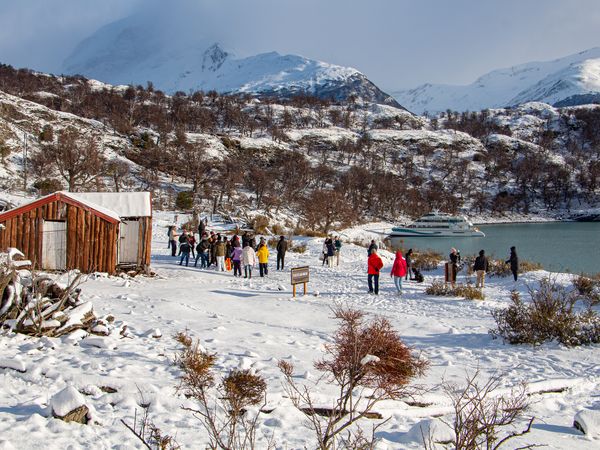 Tourists enjoy a snowy lakeside gathering near Bariloche with snow-capped Patagonian mountains and a cruise ship in the background.