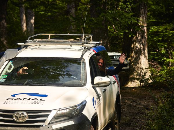 Tourist waving from white Toyota pickup truck on forested road during Patagonia adventure tour.