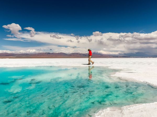 Hiker in red jacket walking across turquoise mineral pools at Salinas Grandes salt flats under dramatic Andean sky.