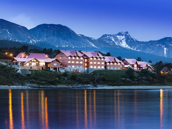 Luxury lodge with pink roof reflects in still waters at dusk, snow-capped Andes mountains beyond, Patagonia.