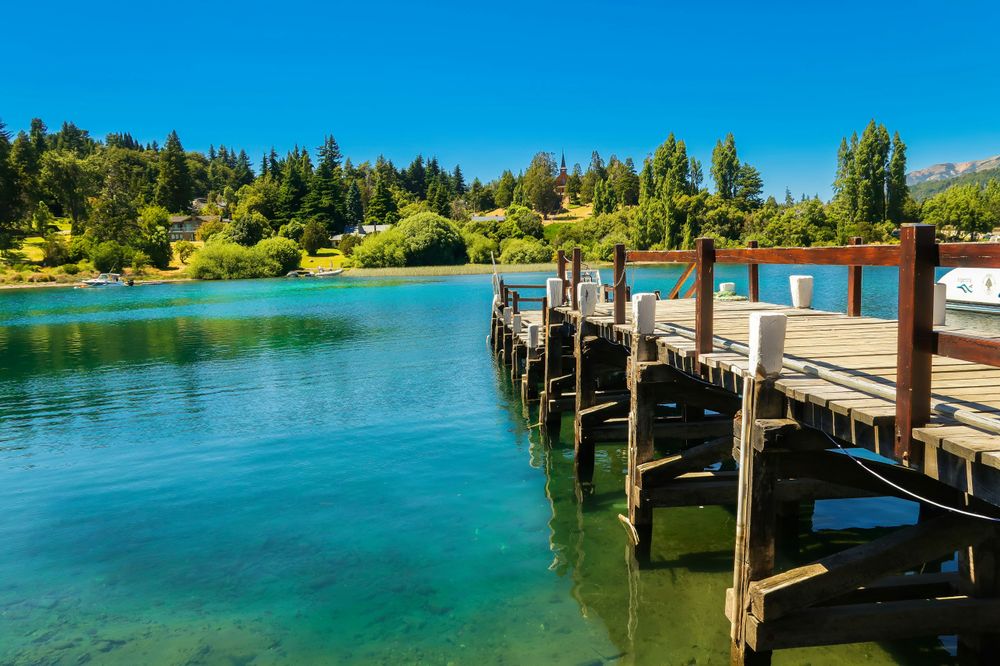 Wooden dock extends into turquoise waters of Lake Nahuel Huapi in Bariloche, with forested shoreline and Andes mountains beyond.