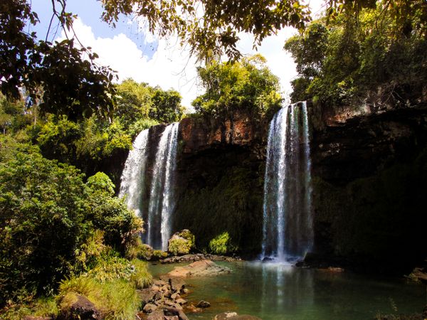Two tall waterfalls plunge into a clear pool, flanked by dense green jungle under a partly cloudy sky.