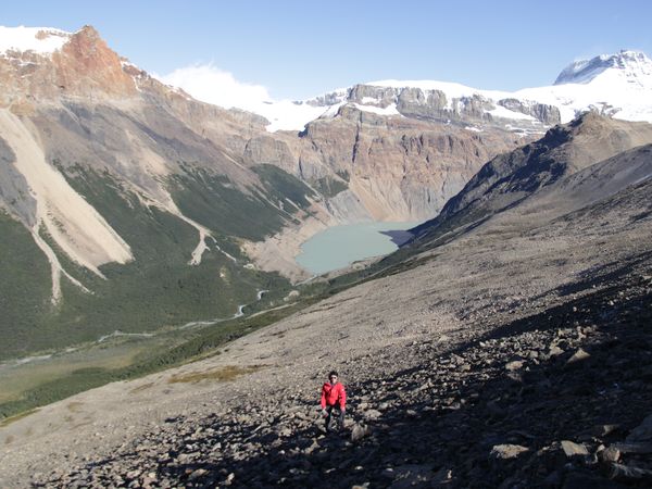 Hiker in red jacket on rocky slope overlooking turquoise glacial lake and snow-capped peaks at Loma del Diablo.