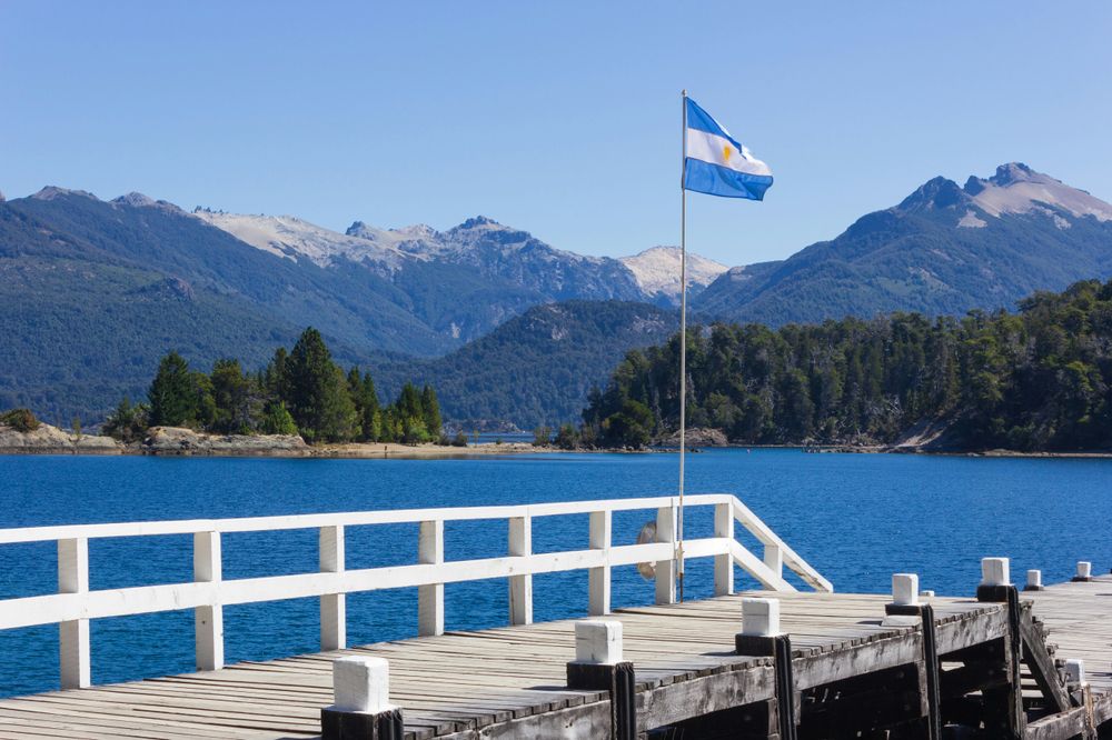 Argentine flag flying from a wooden dock overlooking turquoise Lake Nahuel Huapi with snow-capped Andes mountains in Bariloche.