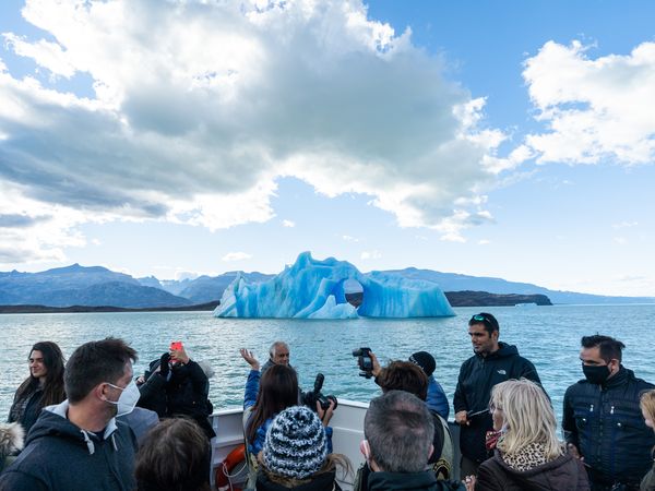 Tourists on a boat observe a striking blue iceberg in the mountainous glacial lake Lago Argentino