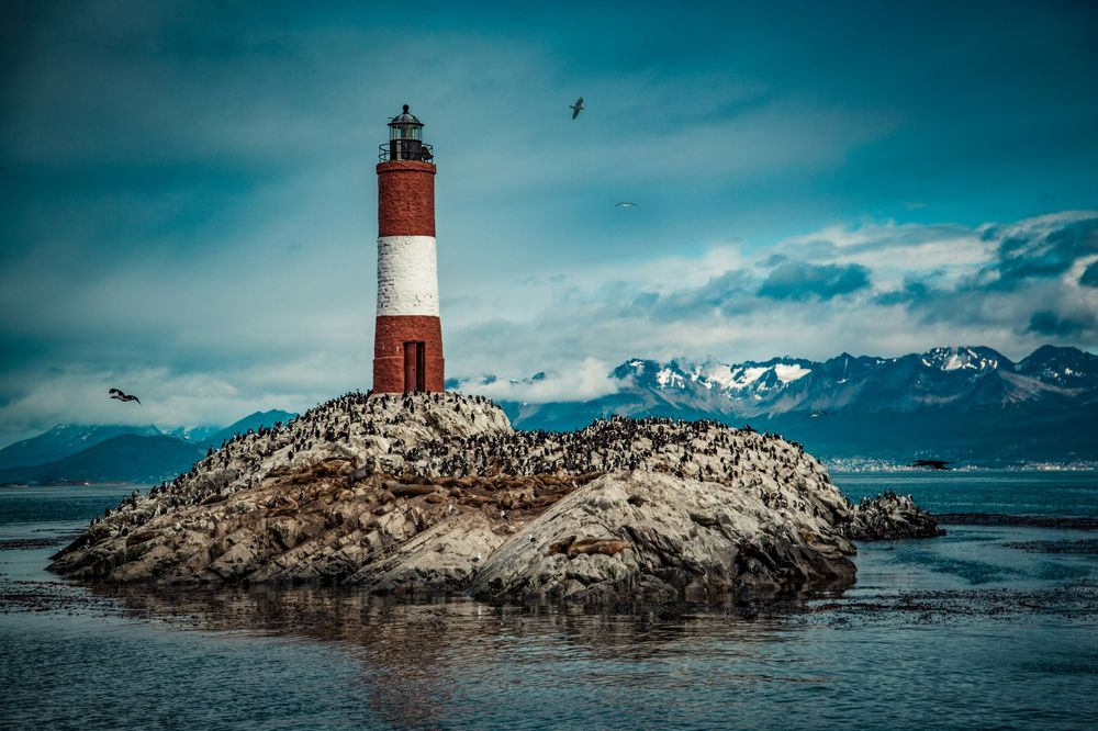 Red and white striped lighthouse on a bird-filled rocky island. Snowy mountains under a cloudy blue sky in the distance.