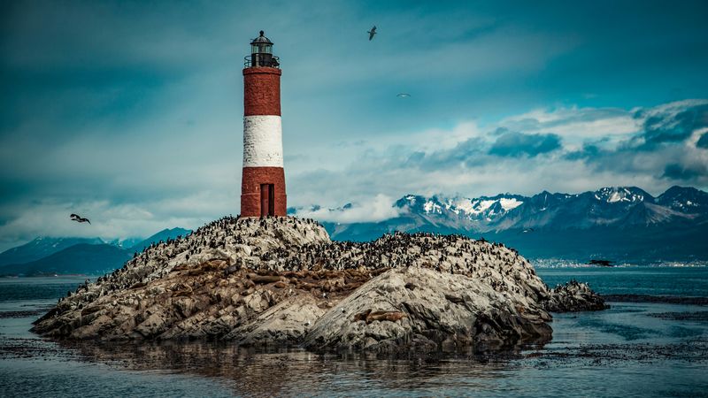 Red and white striped lighthouse on a bird-filled rocky island. Snowy mountains under a cloudy blue sky in the distance.