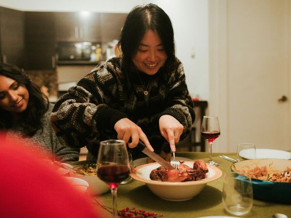 Smiling woman carving roasted meat at a dinner table with wine, as a friend watches.