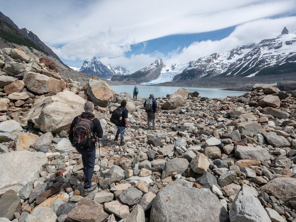 Hikers traverse a rocky moraine toward a turquoise glacial lake surrounded by snow-capped peaks in Patagonia.