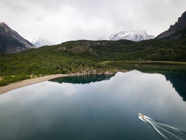 Small boat navigates pristine turquoise waters surrounded by snow-capped mountains and lush green forest in Patagonia.