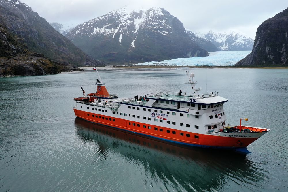 Skorpios cruise ship anchored in a turquoise fjord with snow-capped mountains and Glaciar Guilliard in Patagonia, Chile.