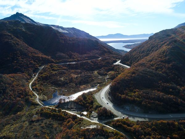 Aerial view of winding roads and rivers through autumn forests in Tierra del Fuego with lakes and mountains visible in the distance.