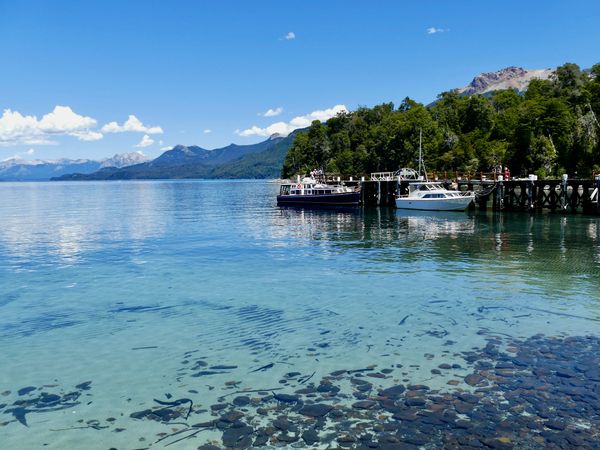 Crystal-clear waters of Lago Nahuel Huapi at Villa La Angostura dock with snow-capped Andes mountains and moored boats.