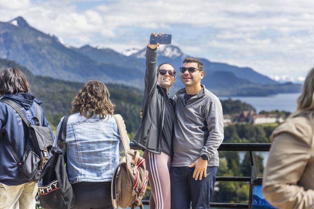 Couple takes selfie at scenic overlook with snow-capped mountains and lake views during Circuito Chico tour near Bariloche.