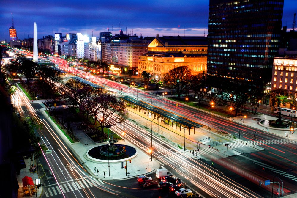 Long exposure of Buenos Aires' 9 de Julio Ave at night, with the Obelisco, streaking car lights, and illuminated buildings.