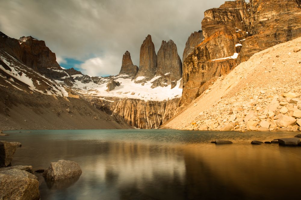 Torres del Paine's iconic granite peaks reflect in turquoise glacial lake waters surrounded by rocky scree and snow.