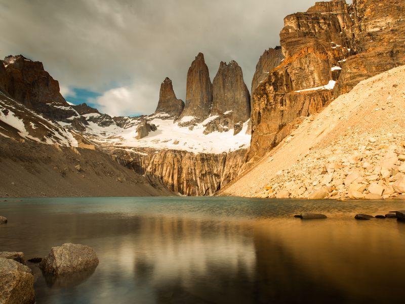 Torres del Paine's iconic granite peaks reflect in turquoise glacial lake waters surrounded by rocky scree and snow.