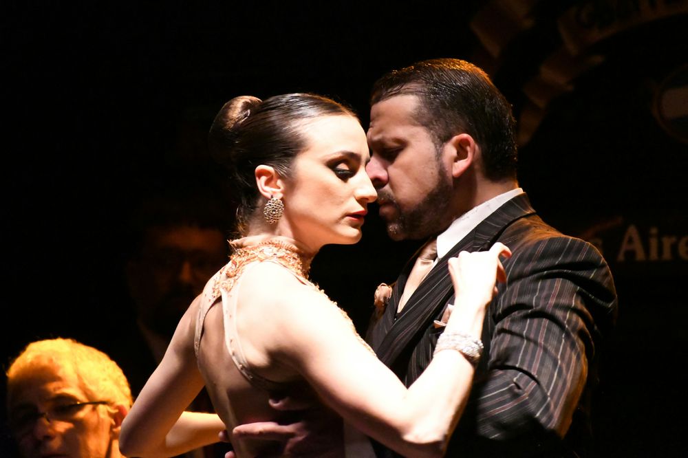 A man in a pinstripe suit and a woman in a beige dress dance tango, close and focused, on a dark stage.