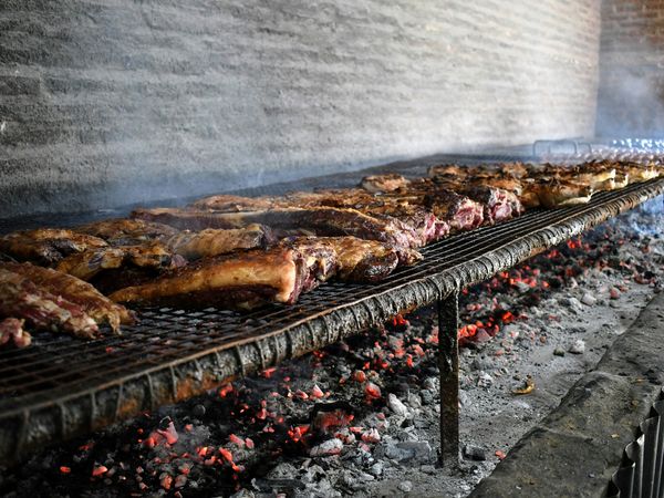 Many thick cuts of meat, likely beef ribs, grilling on a long metal grate over glowing red coals, with smoke rising from a large BBQ.