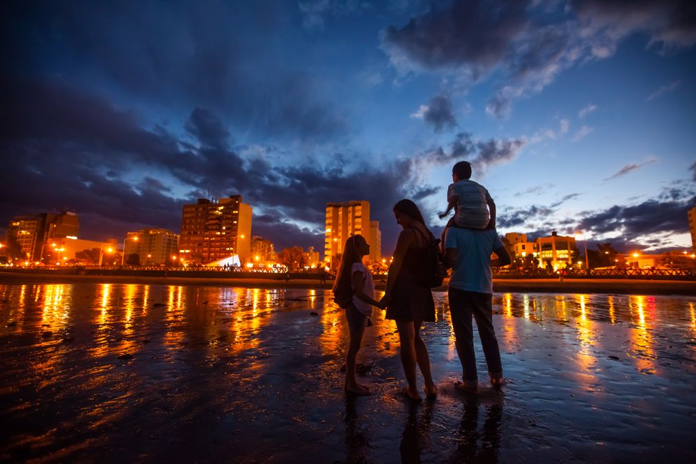 Family wading in the Atlantic Ocean at dusk in Puerto Madryn, with illuminated beachfront buildings reflecting in the water.