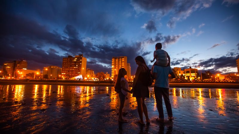 Family wading in the Atlantic Ocean at dusk in Puerto Madryn, with illuminated beachfront buildings reflecting in the water.