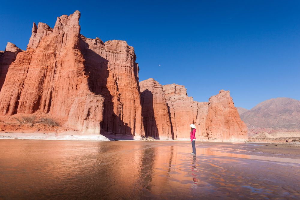 Hiker in pink jacket stands before towering red rock formations reflected in shallow water near Cafayate, Salta.