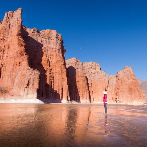 Hiker in pink jacket stands before towering red rock formations reflected in shallow water near Cafayate, Salta.
