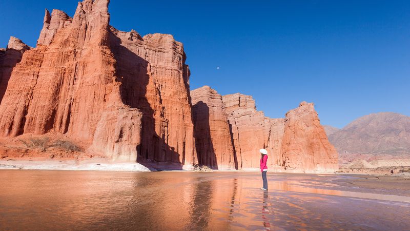 Hiker in pink jacket stands before towering red rock formations reflected in shallow water near Cafayate, Salta.