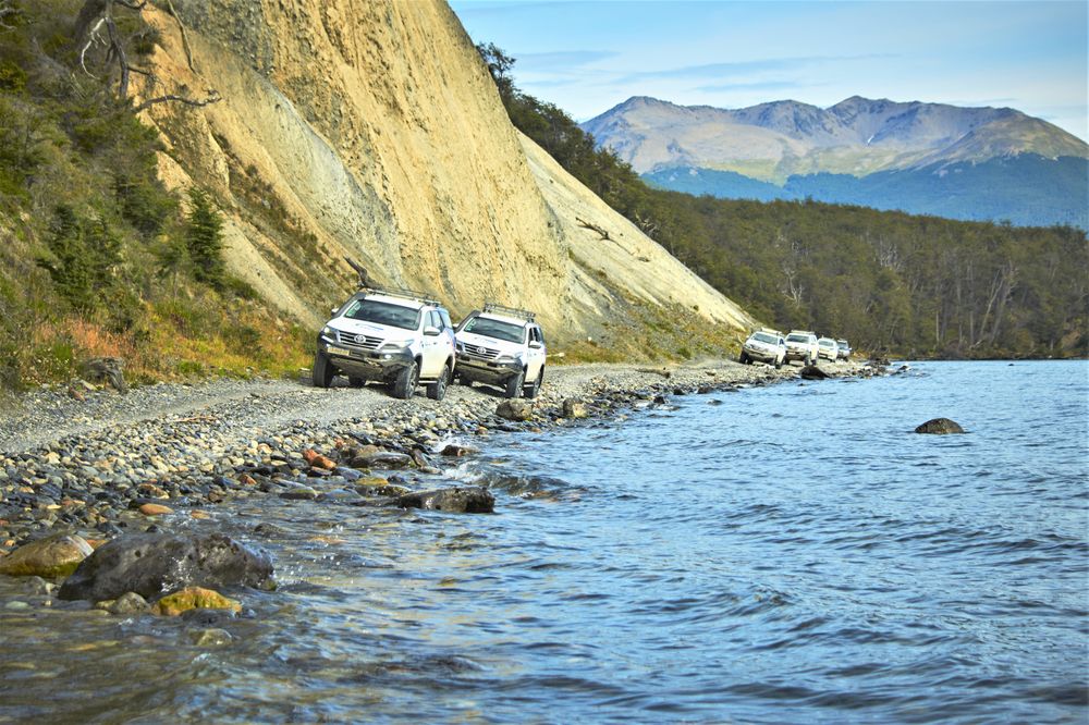 White SUVs parked on a rocky beach beside turquoise waters with a steep cliff and snow-capped mountain in the background near Ushuaia.