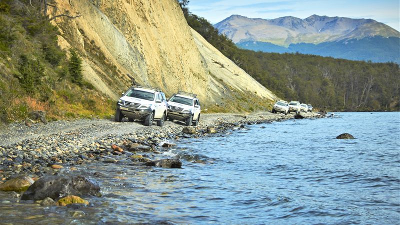White SUVs parked on a rocky beach beside turquoise waters with a steep cliff and snow-capped mountain in the background near Ushuaia.